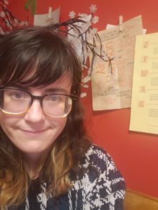 Woman wearing glasses smiling in front of a red wall with broadsides taped up behind her.